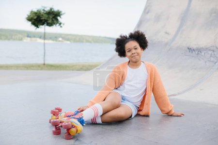 Foto de Linda chica afroamericana patinadora sentada en el parque de la ciudad y sonriendo. - Imagen libre de derechos