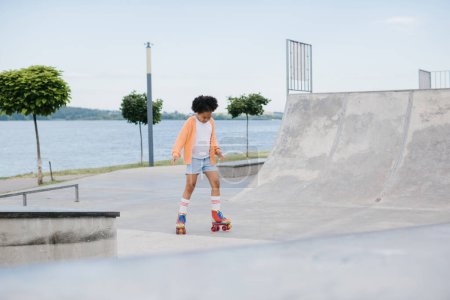 Téléchargez les photos : Petite fille afro-américaine mignonne chevauchant sur patins à roulettes dans un parc d'été. Enfant jouissant de sport en plein air dans le parc, drôle petite fille jouant sur le patin à roulettes sur la route. sport apprentissage concept outdoor - en image libre de droit