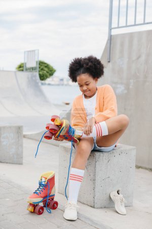Photo for A teenage girl takes off her roller skates after skating. African American girl having fun at the skatepark on the weekend. - Royalty Free Image