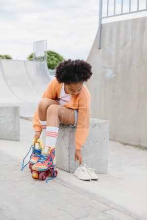 Photo for A teenage girl takes off her roller skates after skating. African American girl having fun at the skatepark on the weekend. - Royalty Free Image