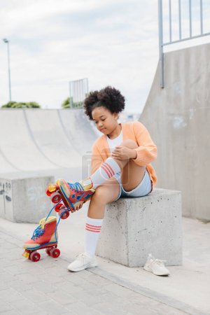 Téléchargez les photos : Une adolescente enlève ses patins à roulettes après avoir patiné. Afro-Américaine s'amuser au skatepark le week-end. - en image libre de droit
