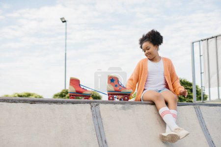 Téléchargez les photos : Adolescente noire africaine active assise en train de se détendre avec ses patins à roulettes au bord de la plage, souriant pendant une journée ensoleillée d'été en vacances, en plein air. Fille joyeuse attrayant contre un ciel bleu. - en image libre de droit