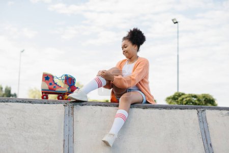 Téléchargez les photos : Adolescente noire africaine active assise en train de se détendre avec ses patins à roulettes au bord de la plage, souriant pendant une journée ensoleillée d'été en vacances, en plein air. Fille joyeuse attrayant contre un ciel bleu. - en image libre de droit