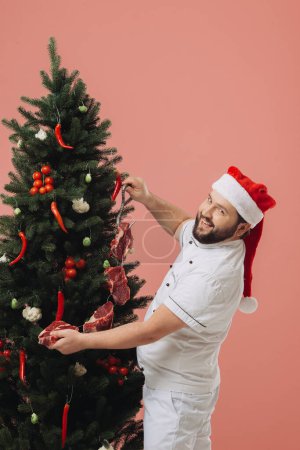 Foto de El concepto de la cocina y las vacaciones de Navidad. Un chef con un sombrero de Santa Claus en el fondo de un árbol de Navidad. - Imagen libre de derechos