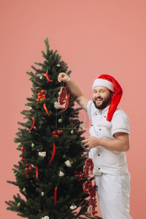 Téléchargez les photos : Le concept de cuisine et vacances de Noël. Un chef dans un chapeau de Père Noël sur le fond d'un arbre de Noël. - en image libre de droit