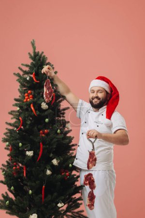 Foto de El concepto de la cocina y las vacaciones de Navidad. Un chef con un sombrero de Santa Claus en el fondo de un árbol de Navidad. - Imagen libre de derechos