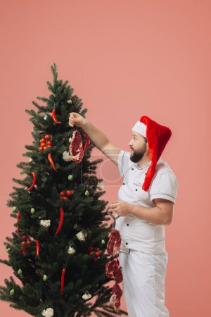 Téléchargez les photos : Le concept de cuisine et vacances de Noël. Un chef dans un chapeau de Père Noël sur le fond d'un arbre de Noël. - en image libre de droit