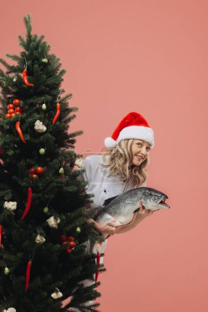 Photo for The concept of cooking and Christmas holidays.  A beautiful woman cook holding a big fish in her hands, against the background of a Christmas tree. - Royalty Free Image