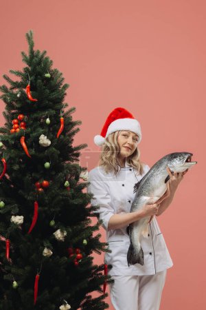 Foto de El concepto de la cocina y las vacaciones de Navidad. Una hermosa cocinera sosteniendo un gran pez en sus manos, sobre el fondo de un árbol de Navidad. - Imagen libre de derechos