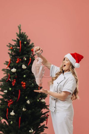 Photo for Concept of cooking and Christmas holidays. A beautiful blonde cook in a Santa hat poses against the background of a Christmas tree. - Royalty Free Image