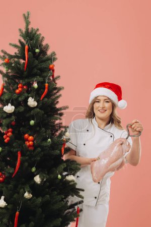Foto de Concepto de cocina y vacaciones de Navidad. Una hermosa cocinera rubia en un sombrero de Santa posa sobre el fondo de un árbol de Navidad. - Imagen libre de derechos