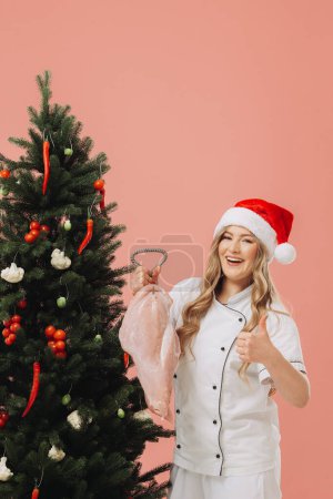Téléchargez les photos : Concept de cuisine et vacances de Noël. Une belle cuisinière blonde dans un chapeau de Père Noël pose sur le fond d'un arbre de Noël. - en image libre de droit