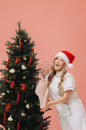 Téléchargez les photos : Concept de cuisine et vacances de Noël. Une belle cuisinière blonde dans un chapeau de Père Noël pose sur le fond d'un arbre de Noël. - en image libre de droit