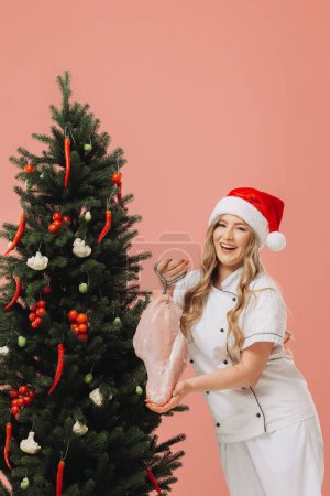 Foto de Concepto de cocina y vacaciones de Navidad. Una hermosa cocinera rubia en un sombrero de Santa posa sobre el fondo de un árbol de Navidad. - Imagen libre de derechos