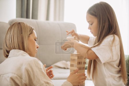 Foto de Una madre feliz y su hija pequeña se divierten jugando un juego de mesa. Madre e hija juegan Jenga en la sala de estar. - Imagen libre de derechos