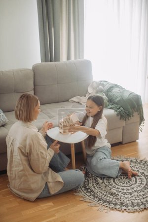 Photo for Young woman and her youthful daughter trying to hold on fallen tower built up of wooden blocks while sitting by table during game - Royalty Free Image