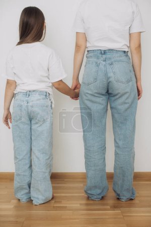 Téléchargez les photos : Mère et petite fille vêtues de jeans et de t-shirts blancs posent contre un mur blanc. - en image libre de droit