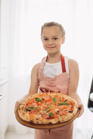 Téléchargez les photos : Une petite cuisinière de 8 ans tient une délicieuse pizza dans une cuisine moderne et légère. - en image libre de droit