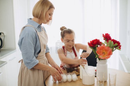 Téléchargez les photos : Une jeune belle mère apprend à sa petite fille à faire de la pâte à crêpes. Mère et fille dans la cuisine ensemble. - en image libre de droit