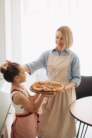 Photo for A beautiful mother and her cute daughter are holding a freshly baked pizza. Concept of cooking at home, - Royalty Free Image