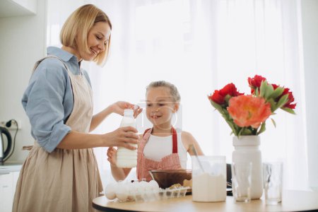 Foto de Una joven y hermosa madre le enseña a su pequeña hija a hacer masa de panqueques. Madre e hija en la cocina juntas. - Imagen libre de derechos
