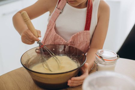 Téléchargez les photos : Une jolie petite fille dans un tablier prépare une pâte à crêpes dans la cuisine. - en image libre de droit
