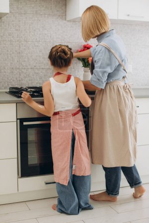 Foto de Una joven madre y su pequeña hija cocinan panqueques juntos para el desayuno. Madre enseña a su hija a cocinar. - Imagen libre de derechos