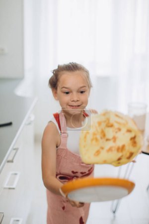 Téléchargez les photos : Une petite fille de 8 ans dans un tablier fait frire des crêpes dans la cuisine. - en image libre de droit