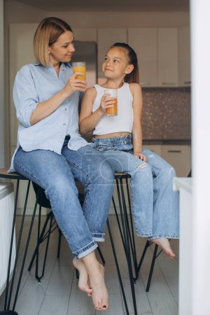 Téléchargez les photos : Mère et fille boivent du jus frais assis dans la cuisine. - en image libre de droit