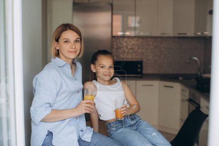 Téléchargez les photos : Mère et fille boivent du jus frais assis dans la cuisine. - en image libre de droit