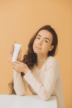 Téléchargez les photos : Une femme tient un tube avec une crème cosmétique. Portrait studio d'une jolie femme sur fond beige. - en image libre de droit
