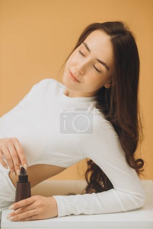 Téléchargez les photos : Une belle brune à la peau propre tient une bouteille de cosmétiques dans ses mains, isolée sur un fond beige. - en image libre de droit