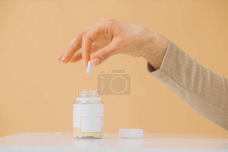 Foto de La mano de una mujer toma una pastilla de una botella. Aislado sobre fondo beige. - Imagen libre de derechos