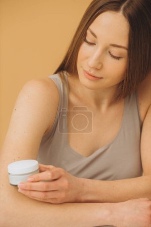 Téléchargez les photos : Portrait de mode d'une belle femme avec une bouteille de crème cosmétique sur fond pastel beige. - en image libre de droit