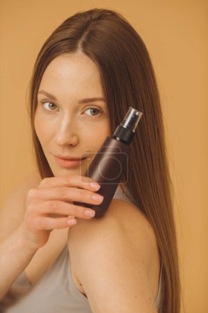 Téléchargez les photos : Portrait de belle femme avec une peau propre et fraîche tenant bouteille de maquillage posant sur fond beige. - en image libre de droit