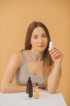 Téléchargez les photos : Belle femme avec des bouteilles de cosmétiques posant sur un fond beige. - en image libre de droit