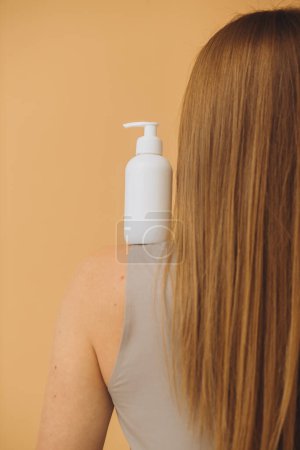 Téléchargez les photos : Une femme tient une bouteille de cosmétiques dans sa main, debout le dos à la caméra. maquette de cosmétiques. - en image libre de droit