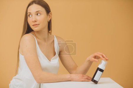 Téléchargez les photos : Portrait de belle femme avec une peau propre et fraîche tenant bouteille de maquillage posant sur fond beige. - en image libre de droit