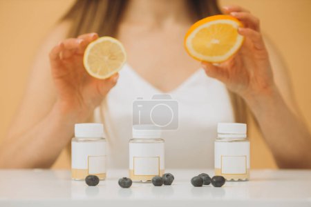 Foto de Hermosa chica con naranja fresca, limón y arándanos y botellas de vitaminas sobre un fondo beige. - Imagen libre de derechos