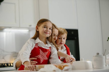 Téléchargez les photos : Deux enfants heureux embrassent leur mère tout en faisant des biscuits de Noël à la maison pendant les vacances d'hiver. Mère heureuse s'amuser avec ses enfants dans la cuisine à la maison. - en image libre de droit