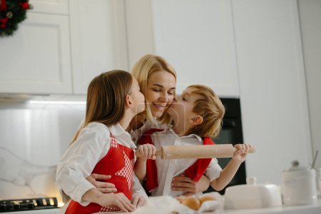 Foto de Dos niños felices abrazan a su madre mientras hacen galletas de Navidad en casa durante las vacaciones de invierno. Feliz madre divirtiéndose con sus hijos en la cocina en casa. - Imagen libre de derechos