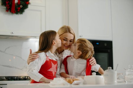 Foto de Dos niños felices abrazan a su madre mientras hacen galletas de Navidad en casa durante las vacaciones de invierno. Feliz madre divirtiéndose con sus hijos en la cocina en casa. - Imagen libre de derechos