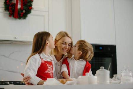 Téléchargez les photos : Deux enfants heureux embrassent leur mère tout en faisant des biscuits de Noël à la maison pendant les vacances d'hiver. Mère heureuse s'amuser avec ses enfants dans la cuisine à la maison. - en image libre de droit