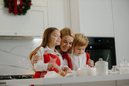 Photo for Two happy children hug their mother while making Christmas cookies at home during winter holidays. Happy mother having fun with her children in the kitchen at home. - Royalty Free Image