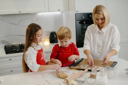 Téléchargez les photos : Jeune mère heureuse avec des enfants qui font des biscuits au pain d'épice de Noël et les mettent sur un plateau dans la cuisine. - en image libre de droit