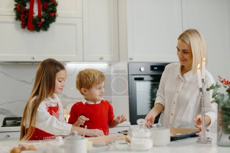 Photo for Young happy mother with children making Christmas gingerbread cookies and putting them on a tray in the kitchen. - Royalty Free Image