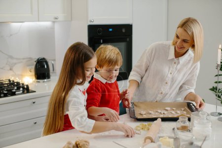 Foto de Joven madre feliz con niños haciendo galletas de jengibre de Navidad y poniéndolas en una bandeja en la cocina. - Imagen libre de derechos