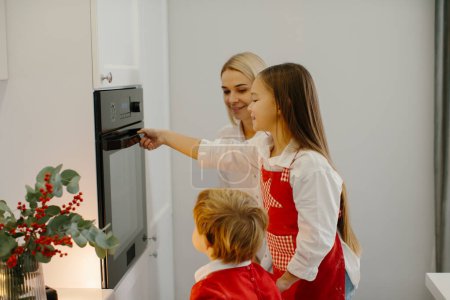 Foto de Madre e hijos hornean pan de jengibre de Navidad en una cocina moderna. Mamá pone una bandeja con galletas en el horno. Preparación de unas vacaciones familiares - Imagen libre de derechos