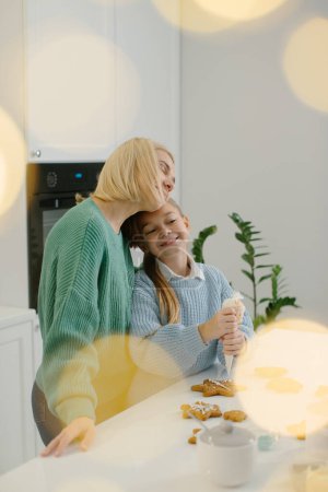 Photo for A mother hugs her little daughter while standing in the kitchen. Mother and daughter decorate gingerbread Christmas cookies with icing. Christmas concept. - Royalty Free Image