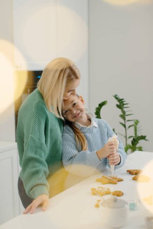 Téléchargez les photos : Une mère étreint sa petite fille alors qu'elle se tient dans la cuisine. Mère et fille décorent les biscuits de Noël en pain d'épice avec du glaçage. Concept de Noël. - en image libre de droit